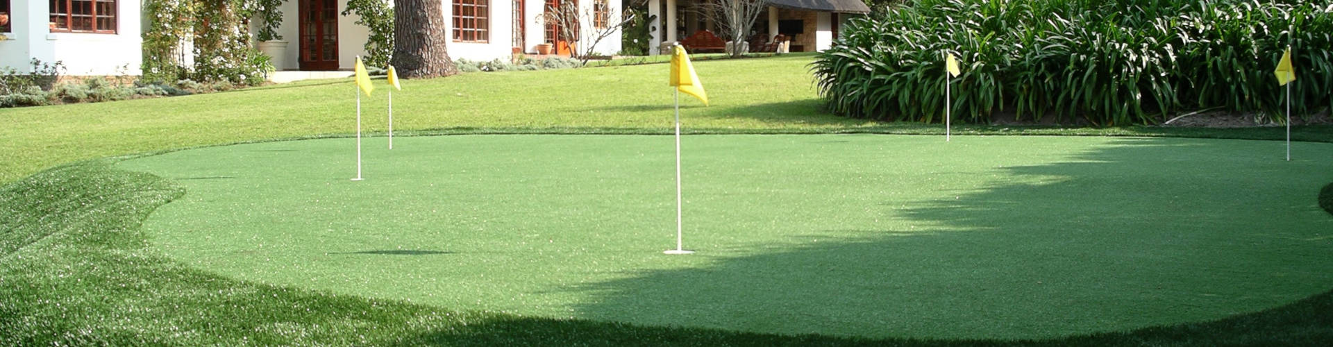 Large backyard putting green with four yellow flag pins surrounded by manicured lawn, tropical garden beds, and a white stucco home with red-framed windows